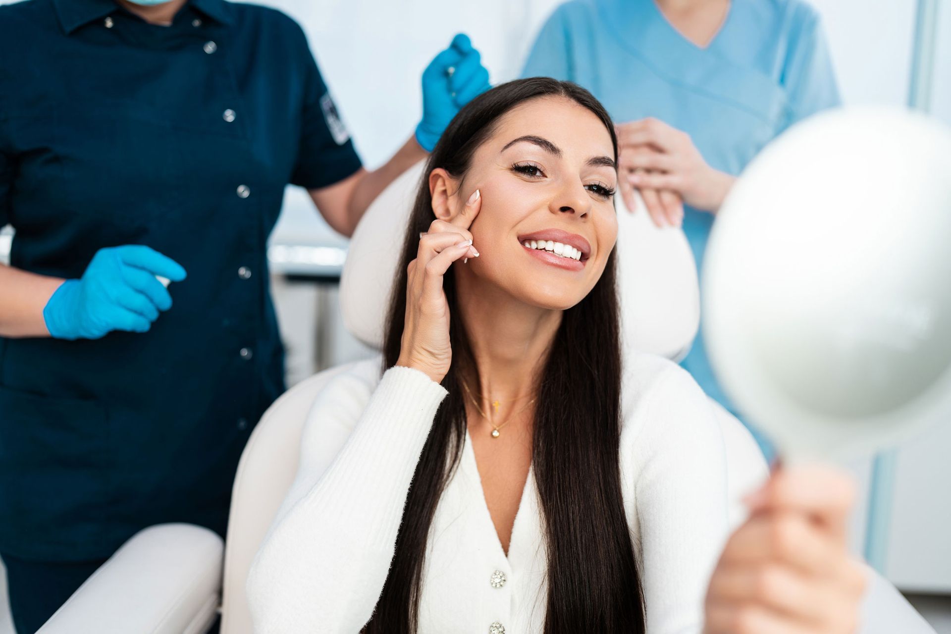 Woman in clinic looking at her face in a mirror, two medical staff in background.