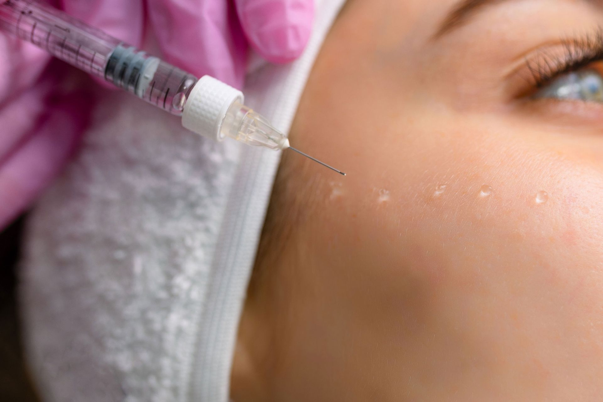 Person receiving a facial injection with a syringe; pink gloves, close-up, white towel, light skin.