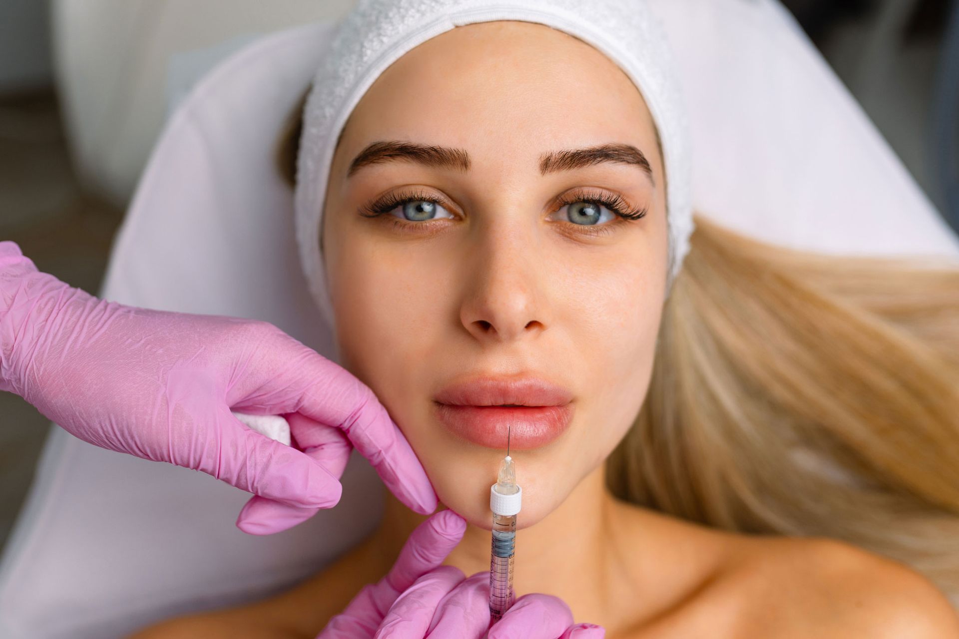 Woman receiving lip filler injections; healthcare professional in pink gloves holds syringe near her lips.