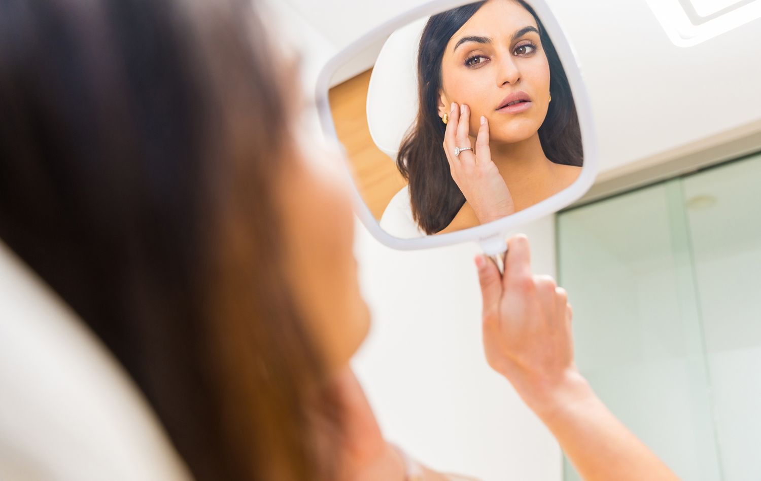 Woman examines her face in a handheld mirror, lit in a bright room.